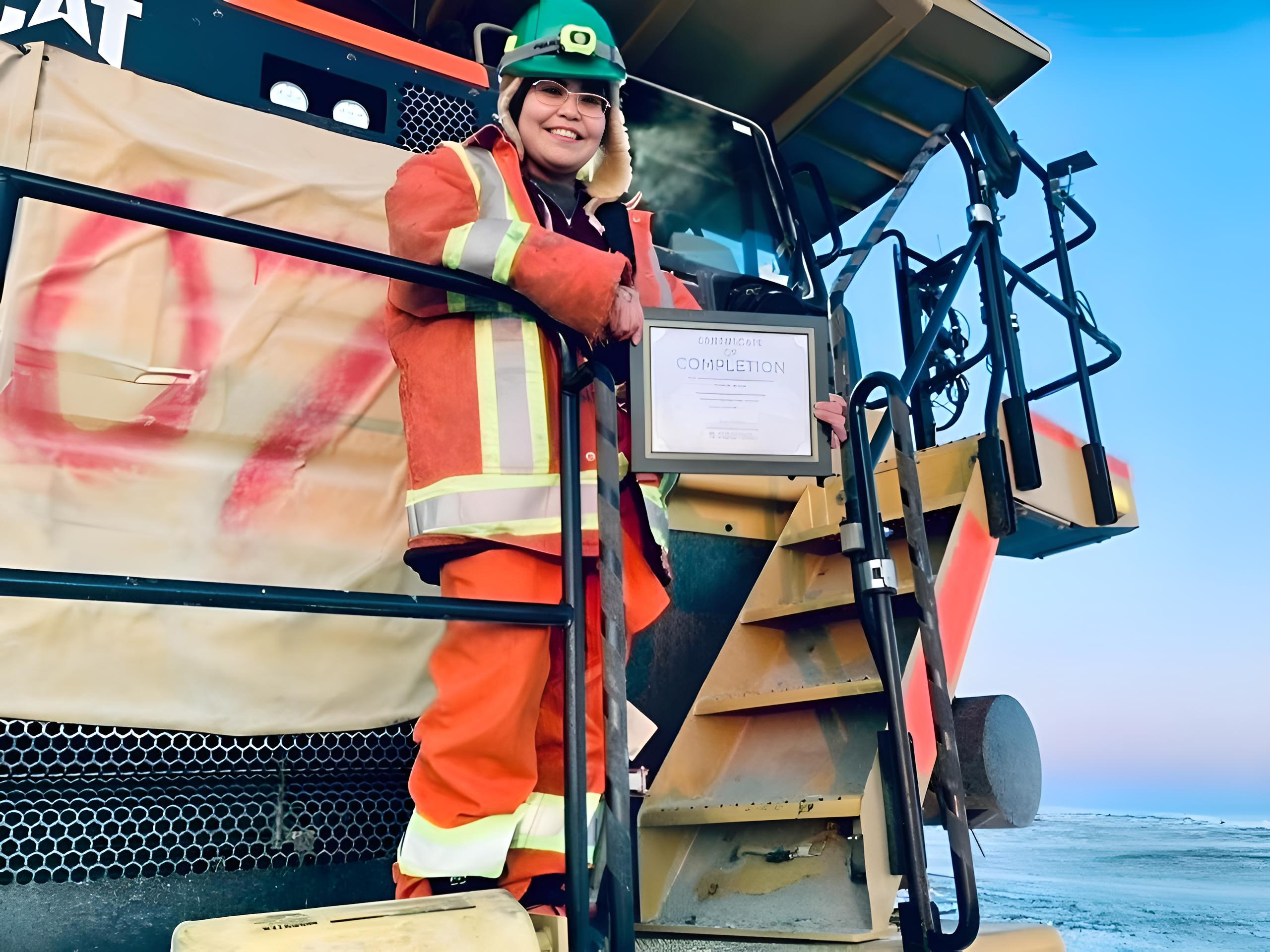 Priscilla Boline standing on the steps of a haul truck at Ekati, holding her Heavy Equipment Operator Certificate of Completion.